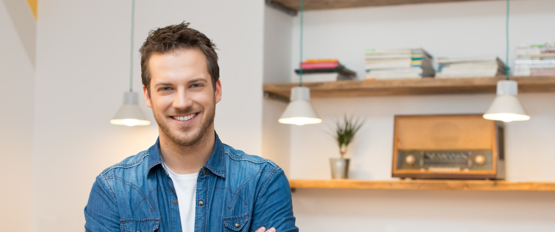 Young man with stubble, wearing a denim shirt, looking relaxed and positive in his apartment.