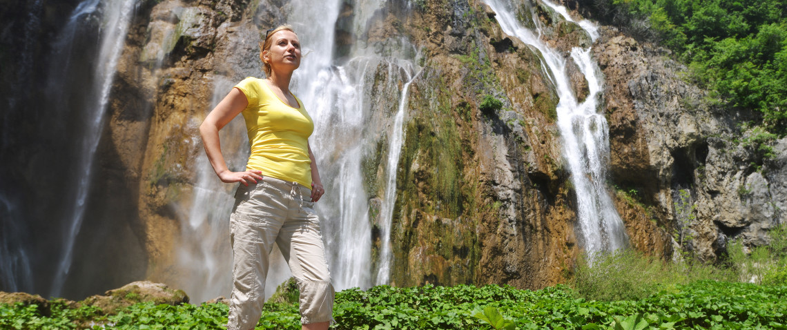 Young fit female in yellow sleeveless top, stood in front of waterfall, after enjoyable walk.