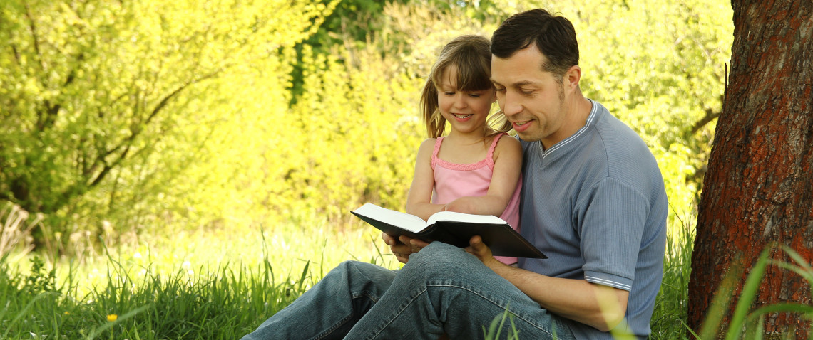 Man reading a book to his young daughter, with his arm around her, sat in the sun beneath a tree.