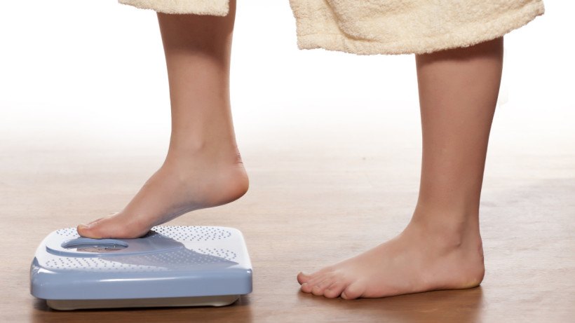 Woman in long bathrobe, barefoot with one foot half way stepping onto weighing scales.