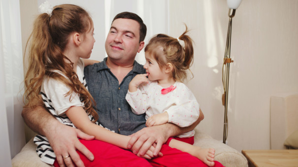 Dad looks lovingly at his two young daughters with pony tails, sat on his knee.